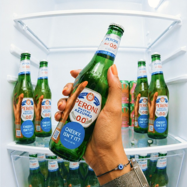 A hand holding a stickered Peroni in front of the fridge
